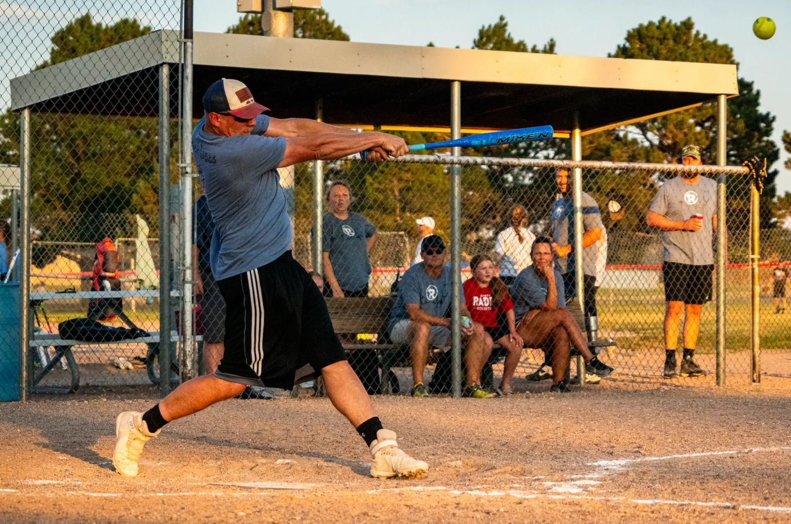 Adult softball batter connecting on a pitch during a The Center Adult Softball League game as teammates and spectators watch from the dugout