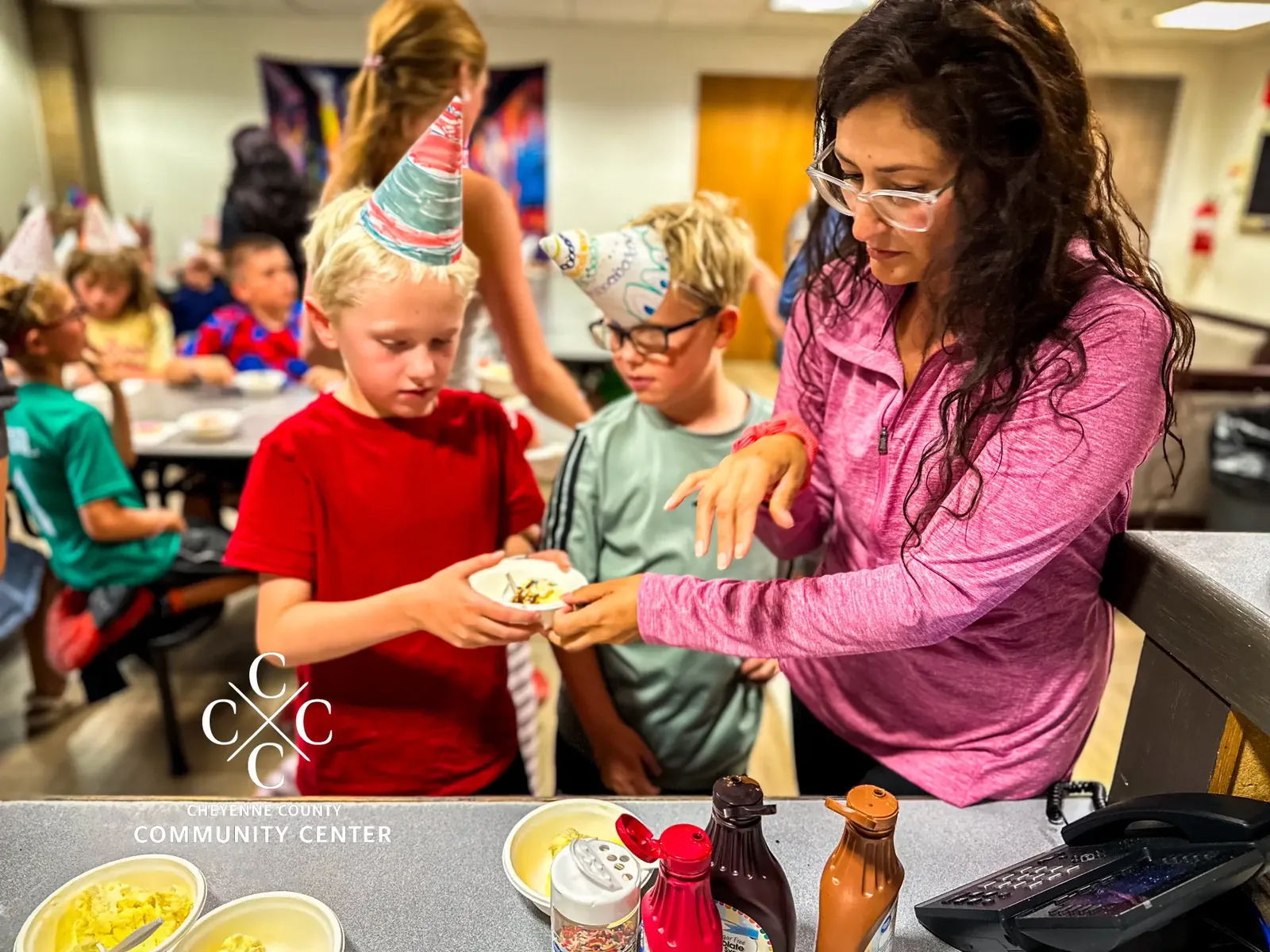 Kids Korner director serving ice cream to two boys wearing handmade birthday party hats while other children eat at tables behind them during the center's 23rd anniversary celebration.