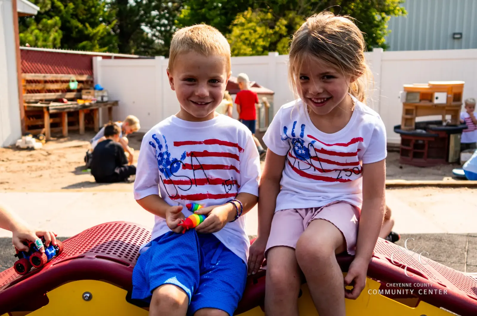 A boy and a girl in handprint-flag USA T-shirts smile while sitting on a red playground climber at Kids Korner.