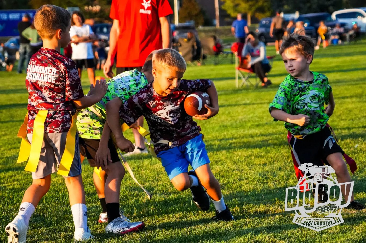 Boy in a maroon flag football jersey carrying the ball past two defenders in yellow flag belts during an evening youth flag football game