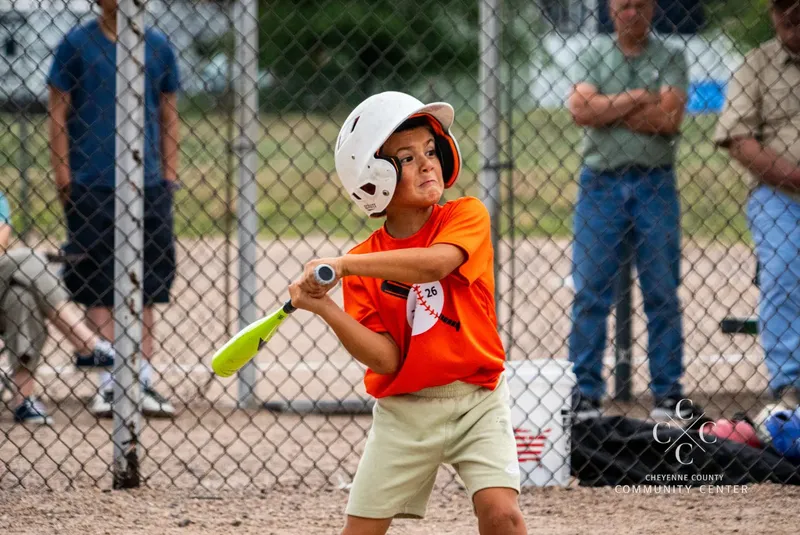Young Rookie League player in an orange Center jersey swinging at the plate during a summer game in Sidney, Nebraska