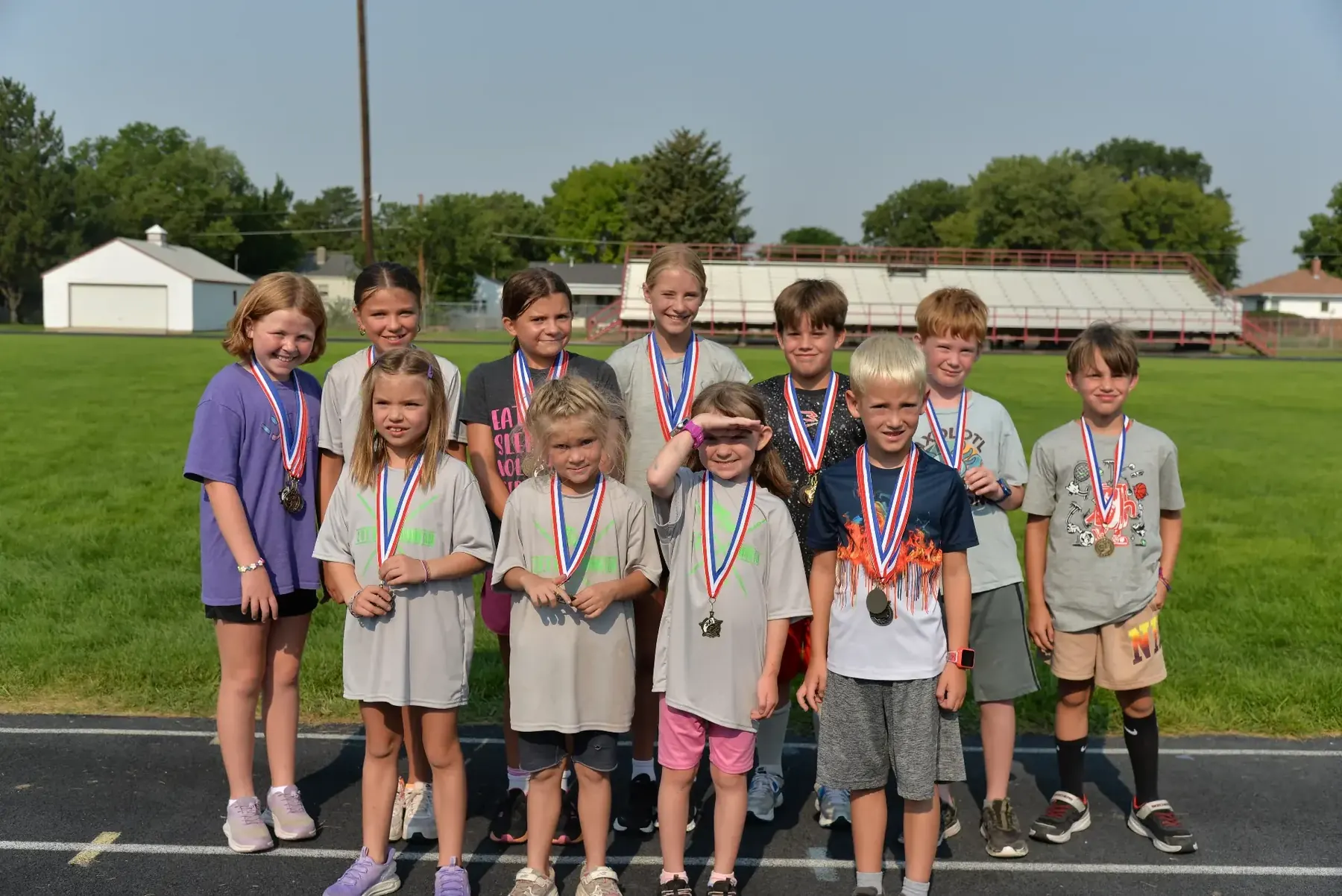 Eleven young runners in Youth Running Club T-shirts stand together on the Weymouth Field track wearing red, white and blue award medals after the season-ending meet.